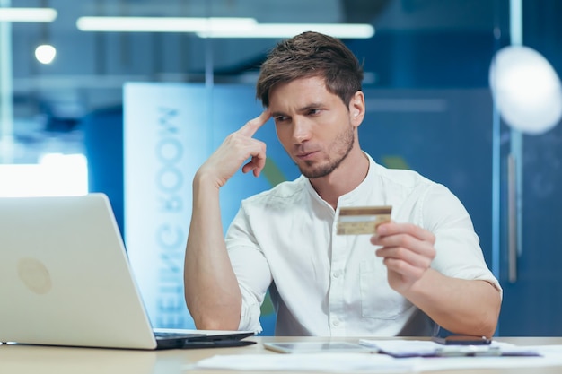 Un jeune homme d'affaires pensif et frustré au bureau essayant de faire un achat dans une boutique en ligne utilise un ordinateur portable contenant une carte de crédit bancaire