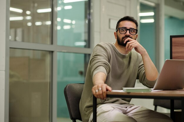 Jeune homme d'affaires pensif à lunettes assis près d'un bureau devant un ordinateur portable