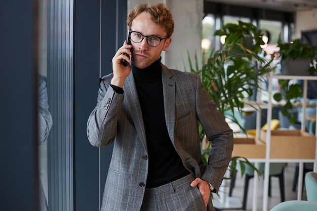 Jeune homme d'affaires pensif portant des lunettes parlant sur un téléphone portable regardant la caméra au bureau
