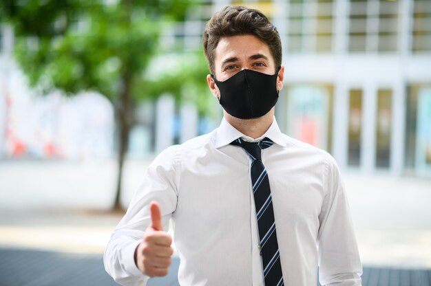 Jeune homme d'affaires en plein air dans un cadre moderne donnant un coup de pouce et portant un masque de protection contre la pandémie de coronavirus covid 19