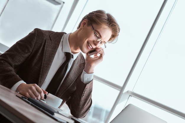 Jeune homme d'affaires portant des lunettes au bureau assis à table avec une tasse de café chaud parlant sur smartphone avec un collègue souriant joyeux