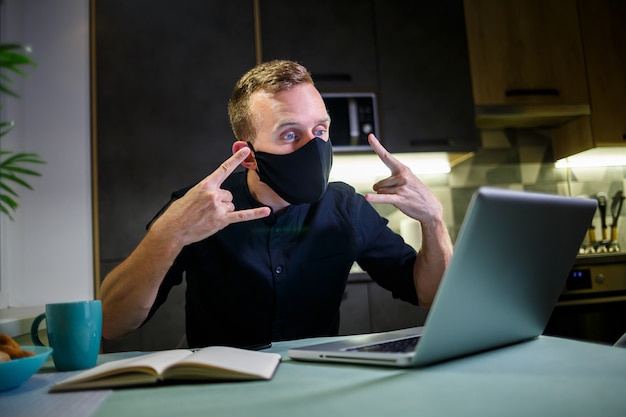 Jeune homme d'affaires portant un masque de protection anti-virus noir, travaillant à la maison, assis à la table de la cuisine avec un ordinateur portable. Travail à domicile en quarantaine