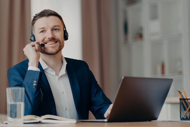 Jeune homme d'affaires positif en costume assis à la table au bureau à domicile portant un casque tout en organisant une conférence en ligne sur un ordinateur portable, un pigiste masculin ayant un appel vidéo avec l'équipe. Concept de travail à distance