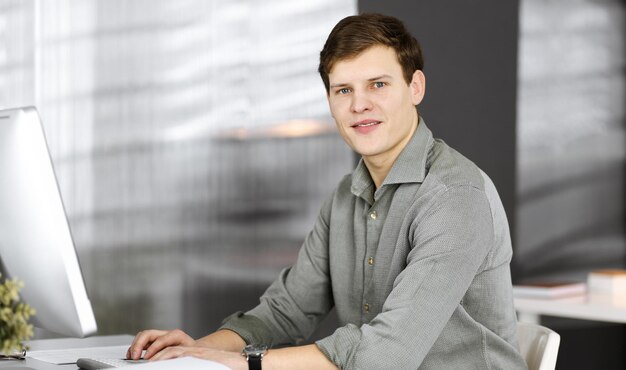 Jeune homme d'affaires et programmeur en chemise verte regarde la caméra, alors qu'il travaille sur son ordinateur, assis au bureau dans une armoire. Headshot ou portrait d'entreprise dans un bureau.