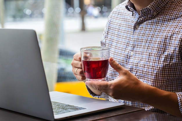 Jeune homme d'affaires prospère assis dans un café, buvant du thé rouge sain et travaillant sur un ordinateur portable. Composition de style de vie avec lumière naturelle