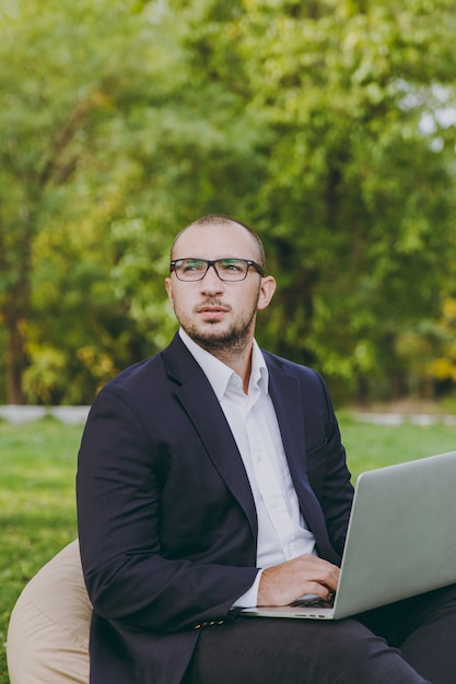 Jeune homme d'affaires prospère en chemise blanche, costume classique, lunettes. L'homme est assis sur un pouf doux, travaillant sur un ordinateur portable dans un parc de la ville sur une pelouse verte à l'extérieur sur la nature. Bureau mobile, concept d'entreprise.