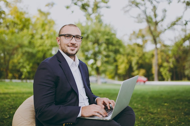 Jeune homme d'affaires prospère en chemise blanche, costume classique, lunettes. L'homme est assis sur un pouf doux, travaillant sur un ordinateur portable dans un parc de la ville sur une pelouse verte à l'extérieur sur la nature. Bureau mobile, concept d'entreprise.
