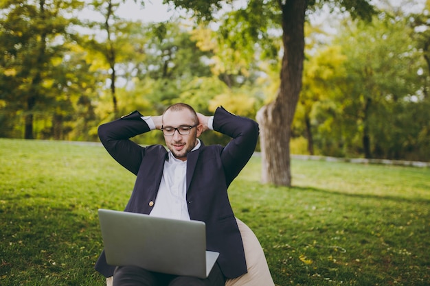 Jeune homme d'affaires prospère en chemise blanche, costume classique, lunettes. L'homme s'assoit sur un pouf doux, se tient les mains derrière la tête, travaille sur un ordinateur portable dans le parc de la ville sur une pelouse verte à l'extérieur. Concept de bureau mobile.