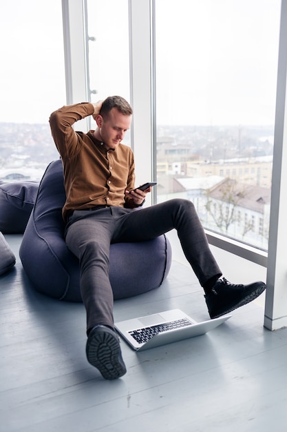 Un jeune homme d'affaires prospère est assis près d'une grande fenêtre du bureau et se détend. Repos après une journée de travail