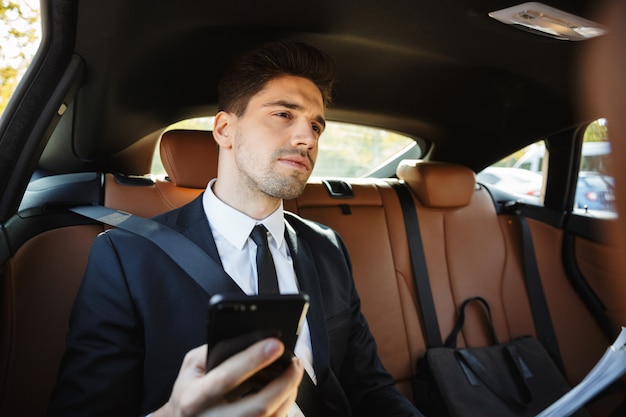 Jeune homme d'affaires prospère de race blanche en costume noir formel tenant un téléphone portable tout en roulant en voiture