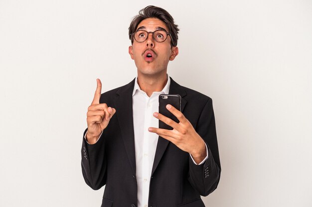 Jeune homme d'affaires de race mixte tenant un téléphone portable isolé sur fond blanc pointant vers le haut avec la bouche ouverte.