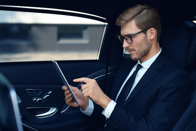 Jeune homme d'affaires réfléchi assis dans la voiture de luxe et utilisant sa tablette