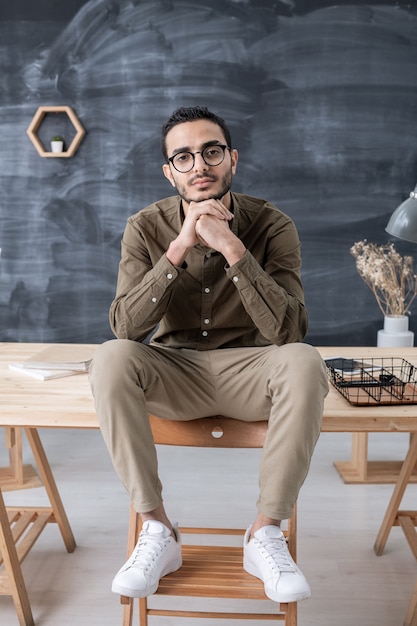 Jeune homme d'affaires reposant dans des vêtements décontractés en vous regardant assis sur une table en bois avec ses pieds sur une chaise devant la caméra