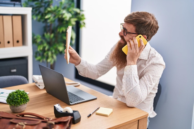 Jeune homme d'affaires rousse parlant sur un smartphone lisant un cahier au bureau
