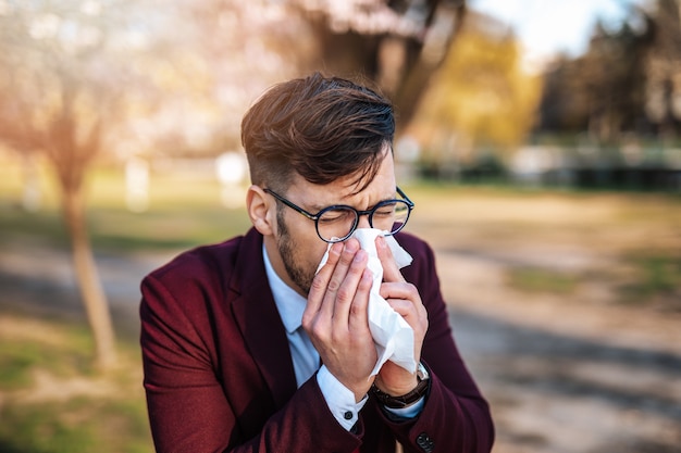 Jeune homme d'affaires se mouchant dans le parc. Allergie, grippe, concept de virus.