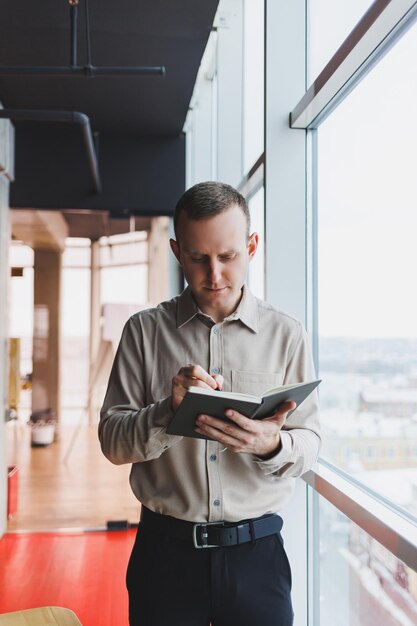 Un jeune homme d'affaires se tient devant une fenêtre panoramique dans un gratte-ciel et prend des notes dans son carnet avec un stylo Un homme en chemise et pantalon d'apparence européenne