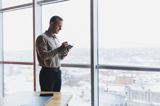 Un jeune homme d'affaires se tient devant une fenêtre panoramique dans un gratte-ciel et prend des notes dans son carnet avec un stylo Un homme en chemise et pantalon d'apparence européenne