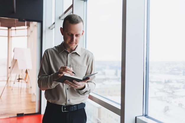 Un jeune homme d'affaires se tient devant une fenêtre panoramique dans un gratte-ciel et prend des notes dans son carnet avec un stylo Un homme en chemise et pantalon d'apparence européenne