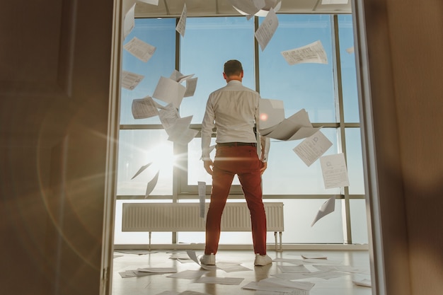Un jeune homme d'affaires se tient le dos en regardant par la fenêtre des documents volent autour
