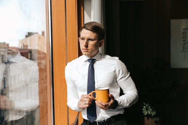 Jeune homme d'affaires se tient avec une tasse de café par la fenêtre de son bureau