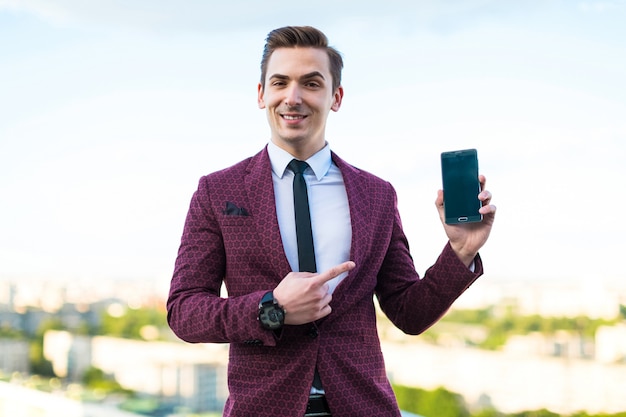 Jeune homme d'affaires sérieux en costume rouge et chemise avec cravate debout sur le toit et spectacle téléphone vide