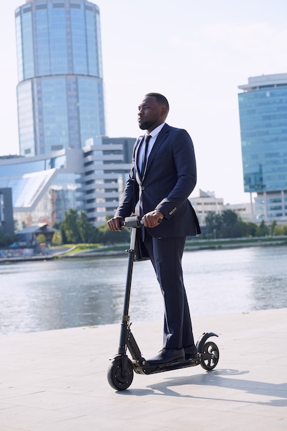 Jeune homme d'affaires sérieux d'origine africaine debout sur un scooter électrique au bord de la rivière contre un groupe de grands bâtiments modernes