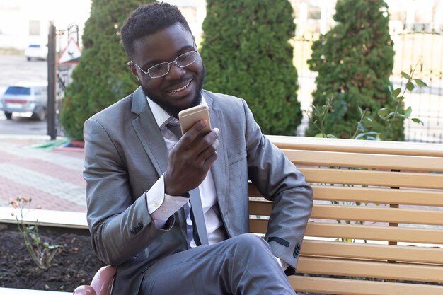 Le jeune homme d'affaires souriant sur le banc de la ville avec téléphone, tablette