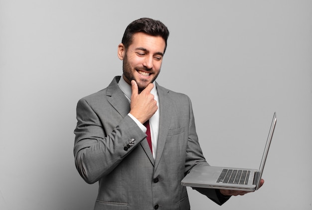 Jeune homme d'affaires souriant avec une expression heureuse et confiante avec la main sur le menton, se demandant et regardant sur le côté et tenant un ordinateur portable