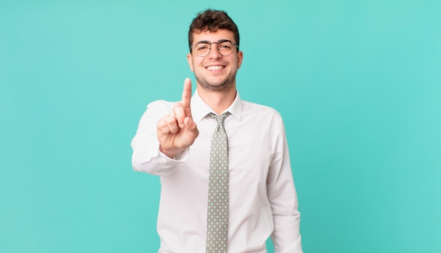 Jeune homme d'affaires souriant fièrement et avec confiance faisant triomphalement la pose numéro un, se sentant comme un leader