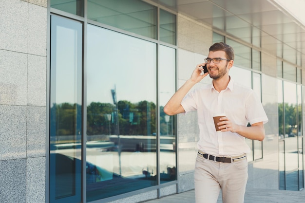 Un jeune homme d'affaires souriant parle au téléphone portable et boit du café tout en marchant près d'un immeuble de bureaux moderne, copiez l'espace. Concept d'entreprise, de communication et de technologie