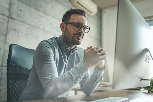 Jeune homme d'affaires souriant et prospère travaillant sur un nouveau projet à l'ordinateur du bureau.