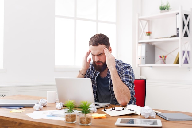 Jeune homme d'affaires stressé, déprimé et malheureux avec un ordinateur portable dans un intérieur de bureau blanc moderne. Bel employé de l'homme au travail avec ordinateur, concept de problèmes de travail. Portrait de style de vie