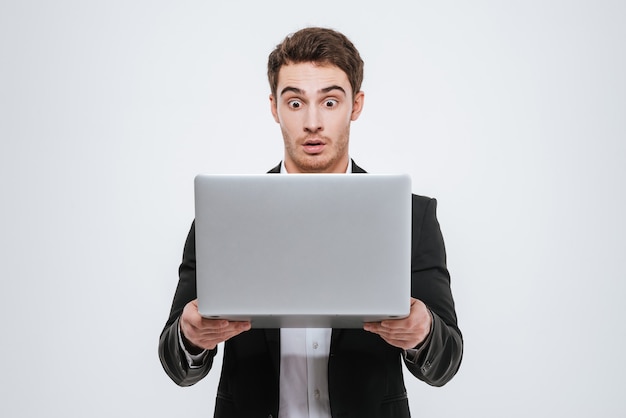 Jeune homme d'affaires surpris en costume noir debout et tenant un ordinateur portable isolé sur un mur blanc