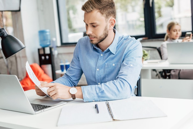 Jeune homme d'affaires tenant des papiers et travaillant avec un ordinateur portable au bureau