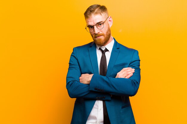 Jeune homme d'affaires à la tête rouge se sentir mécontent et déçu d'avoir l'air sérieux, agacé et fâché avec les bras croisés sur fond orange