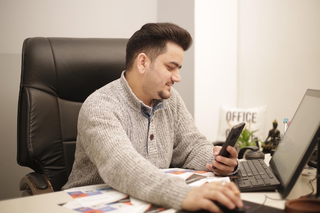 Un jeune homme d'affaires tient un téléphone portable dans une main tout en travaillant sur ordinateur.
