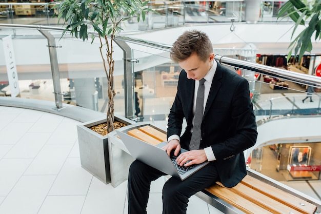 Jeune homme d'affaires travaillant sur ordinateur portable, vêtu d'un costume noir dans un centre commercial, s'assit, à l'intérieur, s'assit
