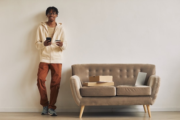 Jeune homme africain appuyé sur le mur à l'aide de son téléphone portable avec ordinateur portable et colis sur le canapé, il reçoit ses commandes