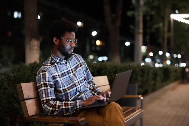 Jeune homme africain assis sur le banc et travaillant en ligne sur un ordinateur portable le soir à l'extérieur