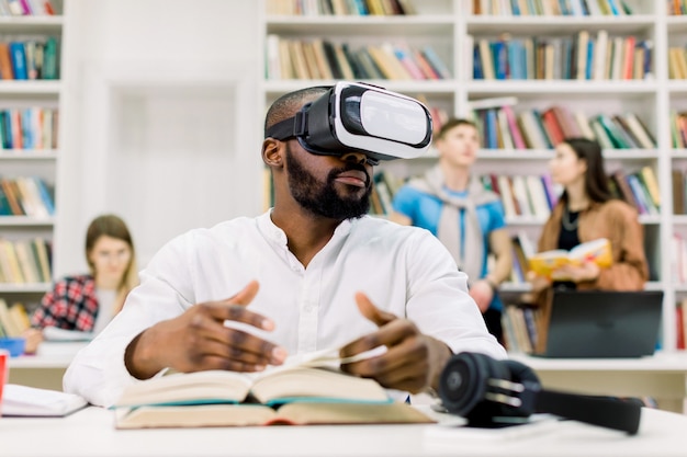 Jeune homme africain attrayant avec barbe portant une chemise blanche décontractée, étudiant en bibliothèque, à l'aide d'un casque de lunettes de réalité virtuelle, feuilletant les pages du livre