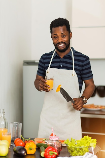 Jeune homme africain barbu joyeux et joyeux tenant un verre de jus de pomme orange fraîchement pressé ou un smoothie et un couteau, préparant une salade de légumes dans la cuisine, souriant en regardant la caméra.