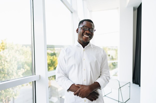 Jeune homme africain en chemise blanche et lunettes posant près d'une grande fenêtre