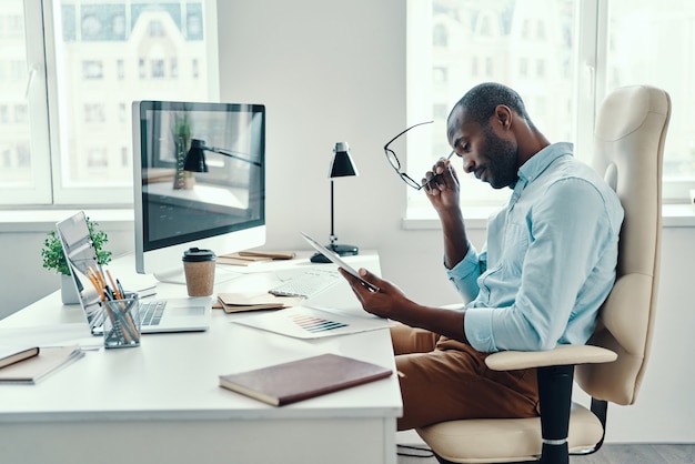 Jeune homme africain concentré en chemise utilisant une tablette numérique tout en travaillant au bureau