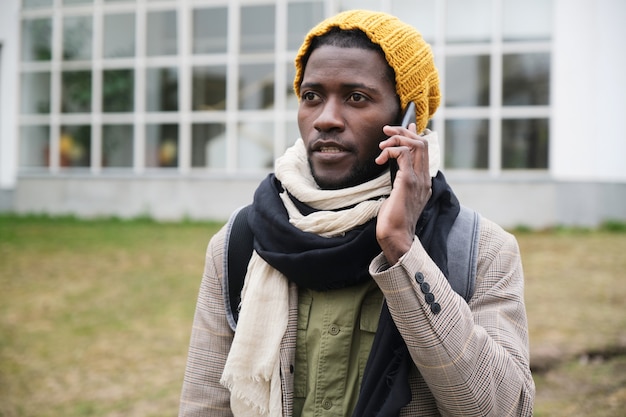 Jeune homme africain dans des vêtements chauds parlant au téléphone portable tout en se tenant à l'extérieur