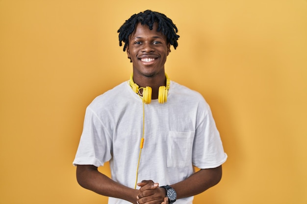 Jeune homme africain avec des dreadlocks debout sur fond jaune avec les mains jointes et les doigts croisés souriant succès détendu et joyeux et optimiste