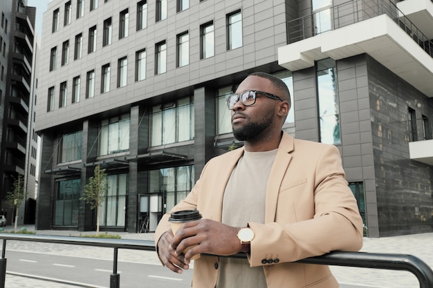 Jeune homme africain à lunettes, boire du café tout en se tenant à l'extérieur, il apprécie la vue sur la ville