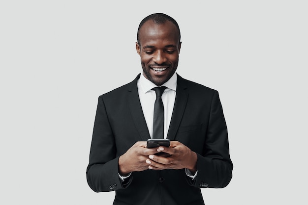 Jeune homme africain moderne en tenue de soirée utilisant un téléphone intelligent et souriant en se tenant debout contre un mur gris