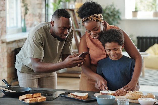 Jeune homme africain avec un smartphone prenant une photo de son mignon petit fils coupant du pain pour des sandwichs tout en préparant le petit-déjeuner pour la famille