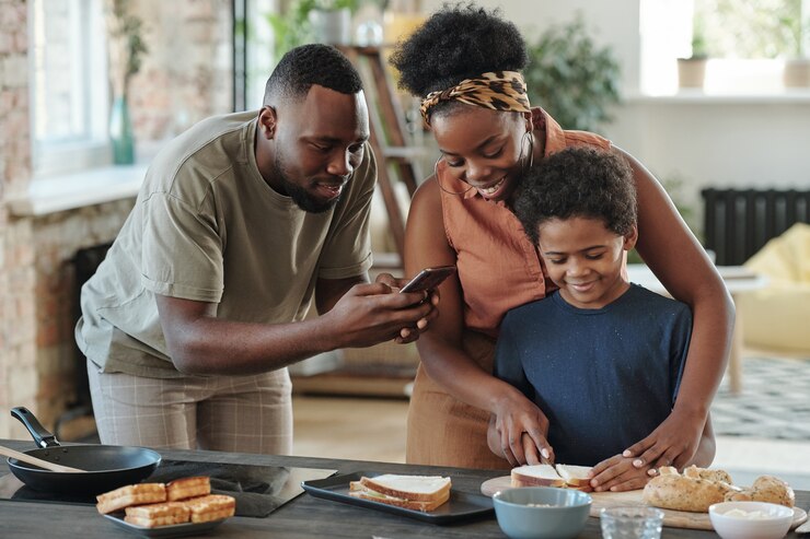 Jeune homme africain avec un smartphone prenant une photo de son mignon petit fils coupant du pain pour des sandwichs tout en préparant le petit-déjeuner pour la famille