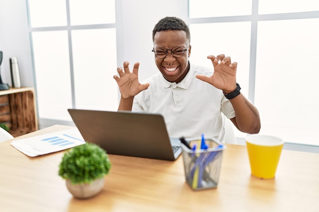 Jeune homme africain travaillant au bureau à l'aide d'un ordinateur portable souriant drôle faisant un geste de griffe comme un chat expression agressive et sexy